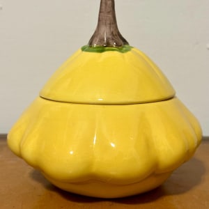 May include: A ceramic squash-shaped container with a lid. The container is a bright yellow color, with a brown stem and green accents. The squash-shaped container is likely used for food storage.