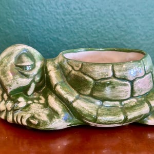 May include: A ceramic turtle-shaped planter featuring a glossy green and white mottled glaze. The turtle has a closed-eye expression and a textured shell that forms the open basin of the planter, with a smooth, light-colored interior finish.