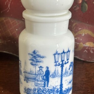 May include: White ceramic jar with a blue and white scene of a couple walking in a park with a street lamp. The jar has a white lid with a rounded top.