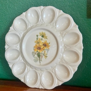 May include: White ceramic egg plate with 12 egg indentations around the edge. The center of the plate features a floral design with yellow flowers. The plate has a scalloped edge and is designed for serving eggs.