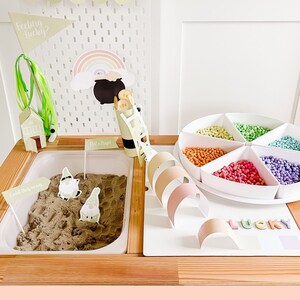 May include: A sensory play setup with a leprechaun trap theme. It includes a wooden table with a sand-filled bin, colorful beads, and decorative elements like a rainbow, a ladder, and signs that read "Feeling Lucky?" and "Not a Trap!"