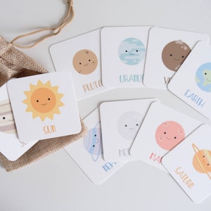 May include: A set of 9 colorful flashcards featuring illustrations of the planets in our solar system, including the sun. Each card has a simple, cartoon-like drawing of a planet with a smiling face and the name of the planet written below. The cards are arranged in a circle on a white surface, with a small burlap bag in the center.