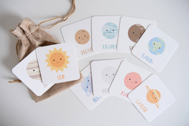 May include: A set of 9 colorful flashcards featuring illustrations of the planets in our solar system, including the sun. Each card has a simple, cartoon-like drawing of a planet with a smiling face and the name of the planet written below. The cards are arranged in a circle on a white surface, with a small burlap bag in the center.