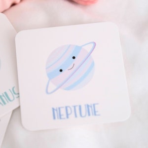 May include: A white card with a blue and purple cartoon planet with a ring around it. The planet has a smiling face. The text "NEPTUNE" is written below the planet.