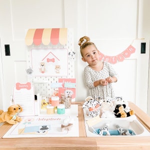 May include: A child plays pretend at a pet adoption booth. The booth is decorated with a pink and yellow awning, a sign that says "Adoption", and a variety of stuffed animals. The child is wearing a white dress with black polka dots and is holding a stuffed animal.