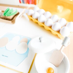 May include: A white toy frying pan with a yellow egg yolk inside. The pan is on a wooden surface next to a blue and white egg carton.