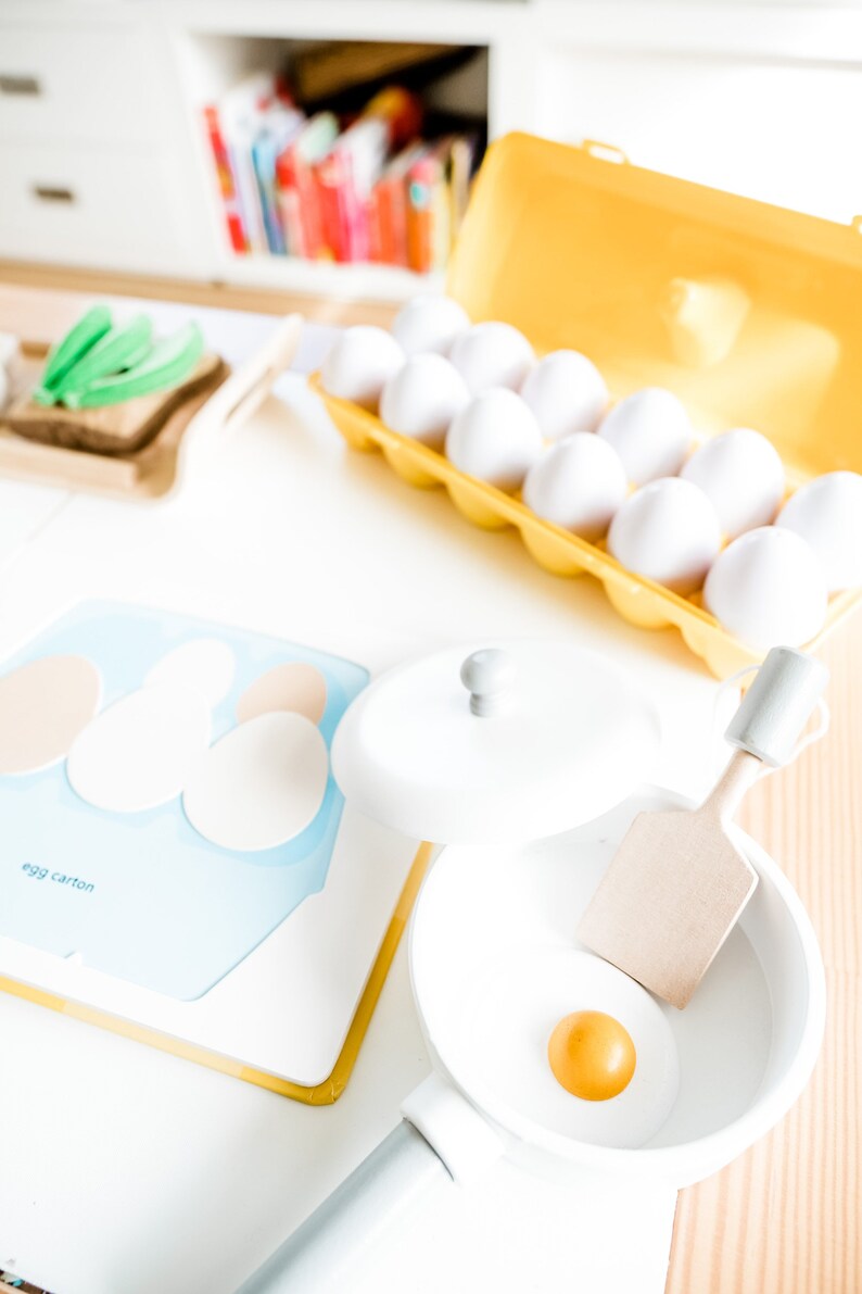 May include: A white toy frying pan with a yellow egg yolk inside. The pan is on a wooden surface next to a blue and white egg carton.