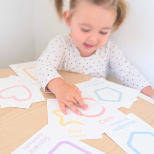 K&ouml;nnte beinhalten: Ein Kind lernt Formen mit bunten Lernkarten. Die Karten zeigen ein Quadrat, einen Kreis, einen Stern, ein Herz und ein Sechseck. Jede Karte tr&auml;gt den Namen der Form in Englisch und Spanisch.