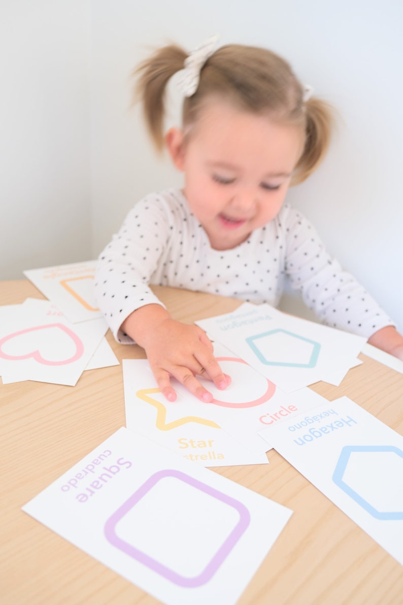 K&ouml;nnte beinhalten: Ein Kind lernt Formen mit bunten Lernkarten. Die Karten zeigen ein Quadrat, einen Kreis, einen Stern, ein Herz und ein Sechseck. Jede Karte tr&auml;gt den Namen der Form in Englisch und Spanisch.
