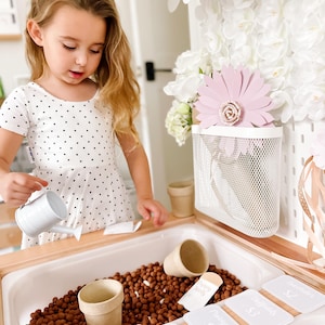 May include: A child engages in imaginative play with a miniature garden. The setup features a white watering can, small plant pots, and price labels for flowers such as tulips and marigolds. The play area is filled with brown, pebble-like material.