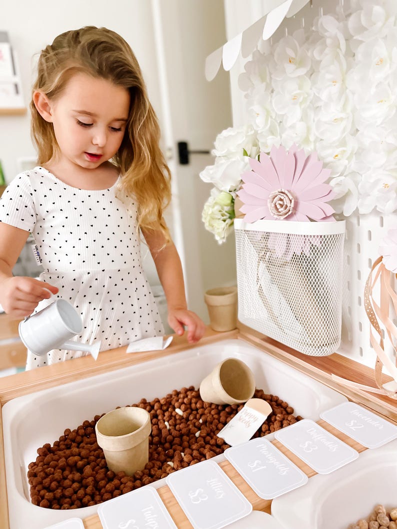 May include: A child engages in imaginative play with a miniature garden. The setup features a white watering can, small plant pots, and price labels for flowers such as tulips and marigolds. The play area is filled with brown, pebble-like material.