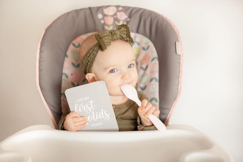 May include: A baby in a high chair wearing a green headband with a bow, holding a card that says "I ate my first solids today" and a pink spoon.