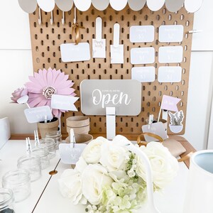 May include: A flower stand with a brown pegboard backdrop and a white awning. The stand displays white roses, paper flowers, and price tags. Signs indicate prices for bouquets, poppies, and other flowers. The sign reads "Open."