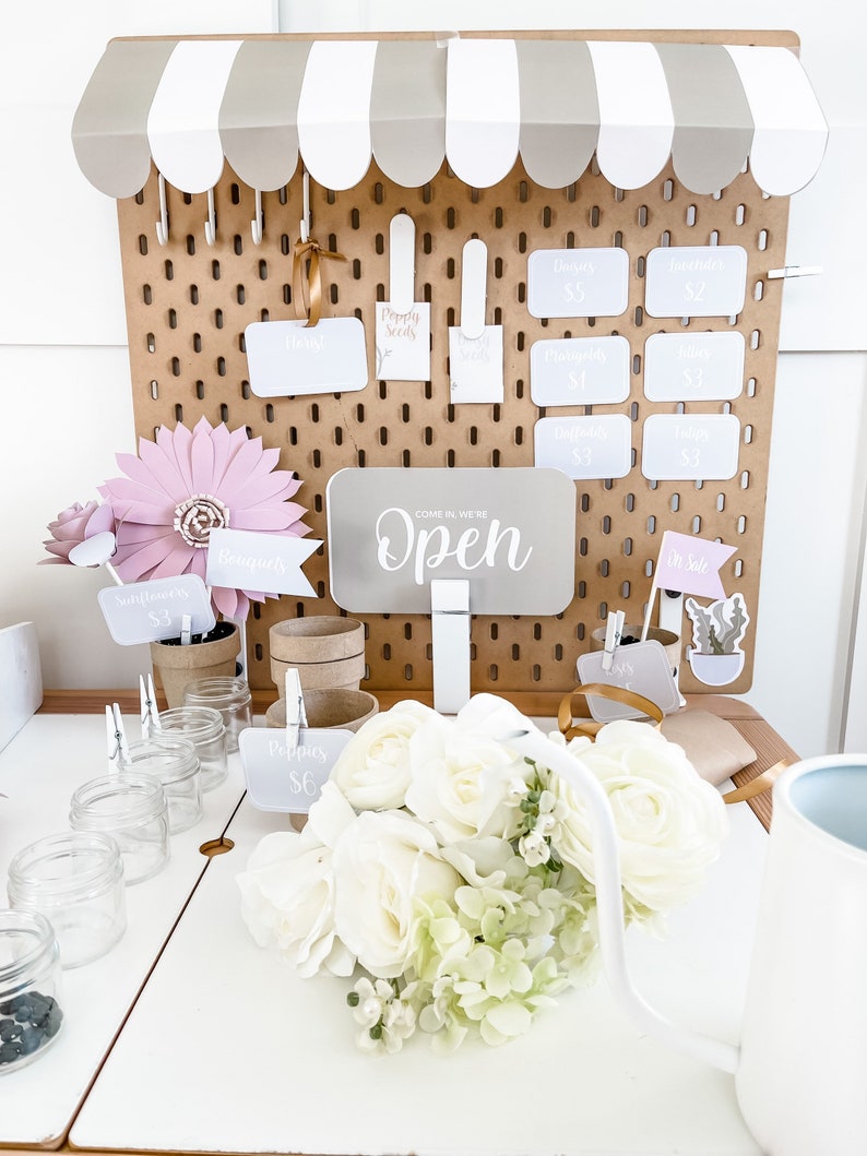 May include: A flower stand with a brown pegboard backdrop and a white awning. The stand displays white roses, paper flowers, and price tags. Signs indicate prices for bouquets, poppies, and other flowers. The sign reads "Open."