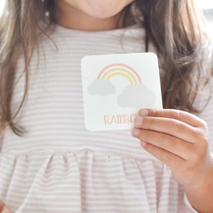 May include: A child holds a flashcard with a rainbow and clouds illustration and the word "Rainbow" printed on it. The card is part of a memory game set by Goodnight Fox.
