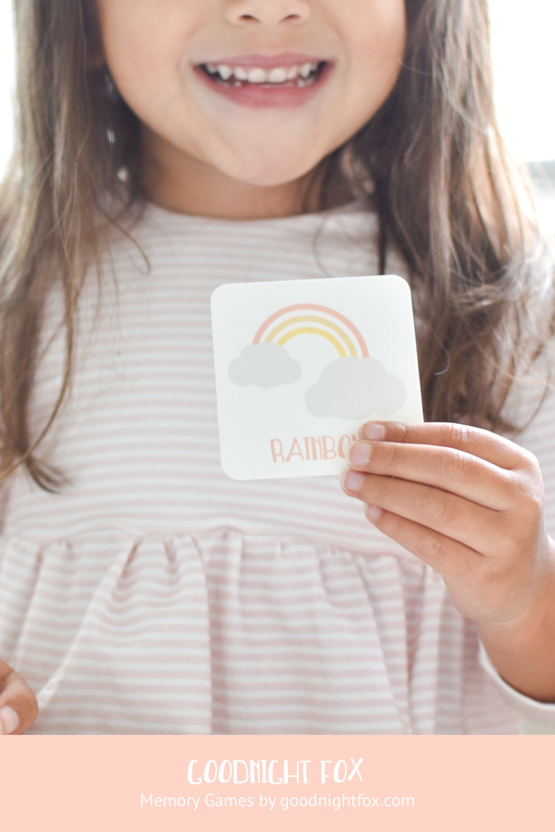 May include: A child holds a flashcard with a rainbow and clouds illustration and the word "Rainbow" printed on it. The card is part of a memory game set by Goodnight Fox.