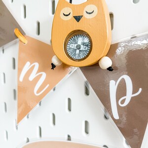 May include: A wooden owl-shaped compass with a black string attached. The compass has a black face with white markings and a black needle. The owl is painted a light brown color with black eyes and a black beak. The owl is hanging on a white pegboard with brown felt pennants that say "m" and "p" hanging below.