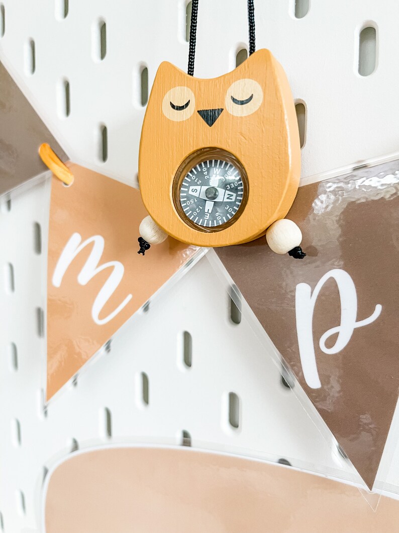 May include: A wooden owl-shaped compass with a black string attached. The compass has a black face with white markings and a black needle. The owl is painted a light brown color with black eyes and a black beak. The owl is hanging on a white pegboard with brown felt pennants that say "m" and "p" hanging below.