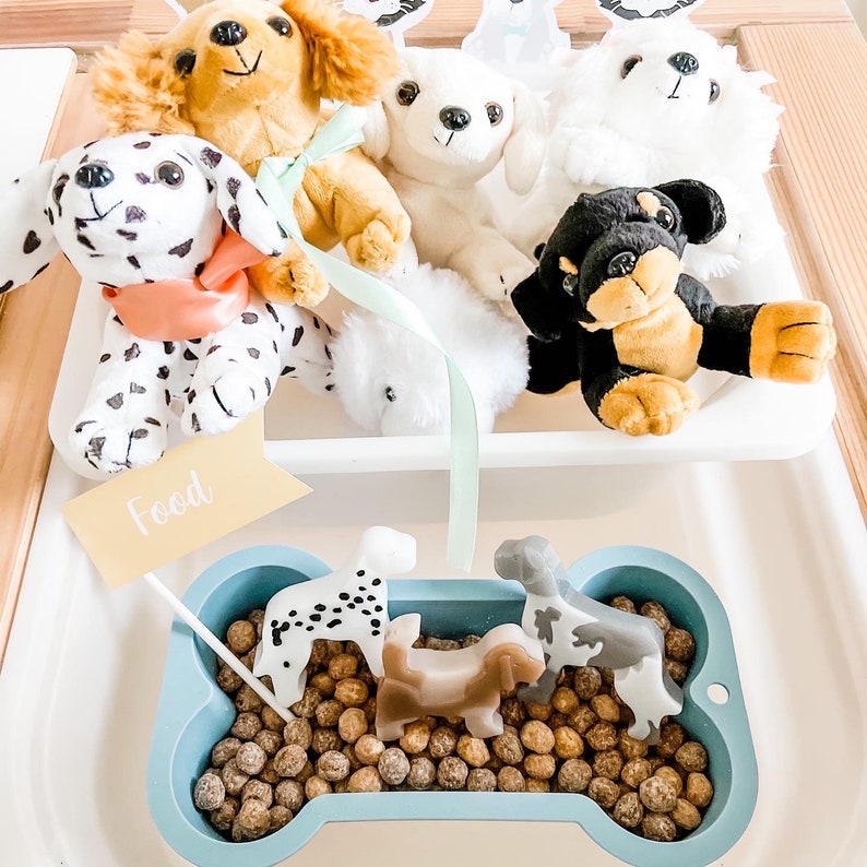 May include: A blue bone-shaped dish filled with brown dog food kibble.  Five small plastic toy dogs are in the dish, including a black and white spotted dog, a brown dog, and a gray and white spotted dog.  The word "Food" is written on a yellow sign.