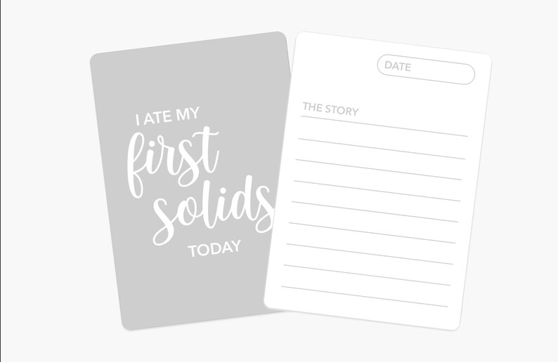 May include: A gray card with the text "I ate my first solids today" in white. A white card with lines for writing a date and a story is attached to the gray card.