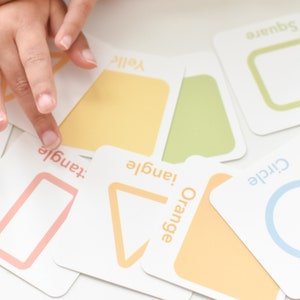 May include: A set of colorful flash cards featuring different shapes and colors. The cards are labeled with the shape name and color, such as "Orange Triangle" and "Yellow Circle". The cards are designed for children ages 2 and up.