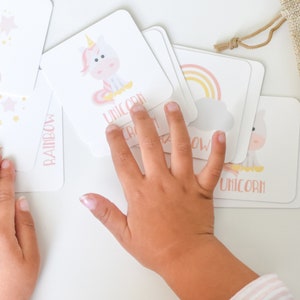May include: A set of colorful memory game cards featuring illustrations of a unicorn, rainbow, and stars. The cards are laid out on a white surface with a child's hand reaching for one of the cards. The text "GOODNIGHT FOX" and "Memory Games by goodnightfox.com" is printed on the bottom of the image.