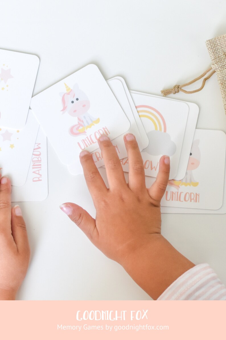 May include: A set of colorful memory game cards featuring illustrations of a unicorn, rainbow, and stars. The cards are laid out on a white surface with a child's hand reaching for one of the cards. The text "GOODNIGHT FOX" and "Memory Games by goodnightfox.com" is printed on the bottom of the image.