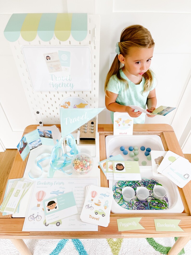 May include: A wooden playset with a travel agency theme. The playset includes a cash register, a booking form, a passport station, and a luggage station. The playset is decorated with travel-themed images and text, such as "Travel Agency", "Booking Form", "Passports", and "Luggage".