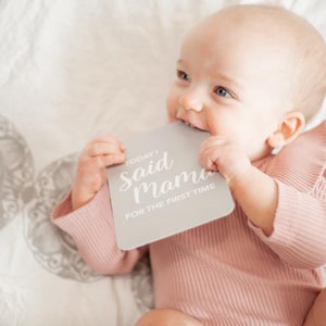 May include: A baby in a pink ruffled onesie holds a gray milestone card that reads "Today I Said Mama for the First Time."
