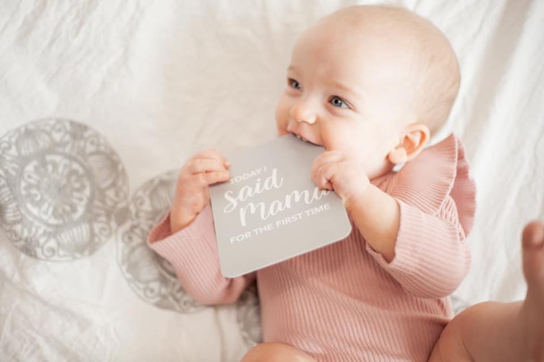 May include: A baby in a pink ruffled onesie holds a gray milestone card that reads "Today I Said Mama for the First Time."