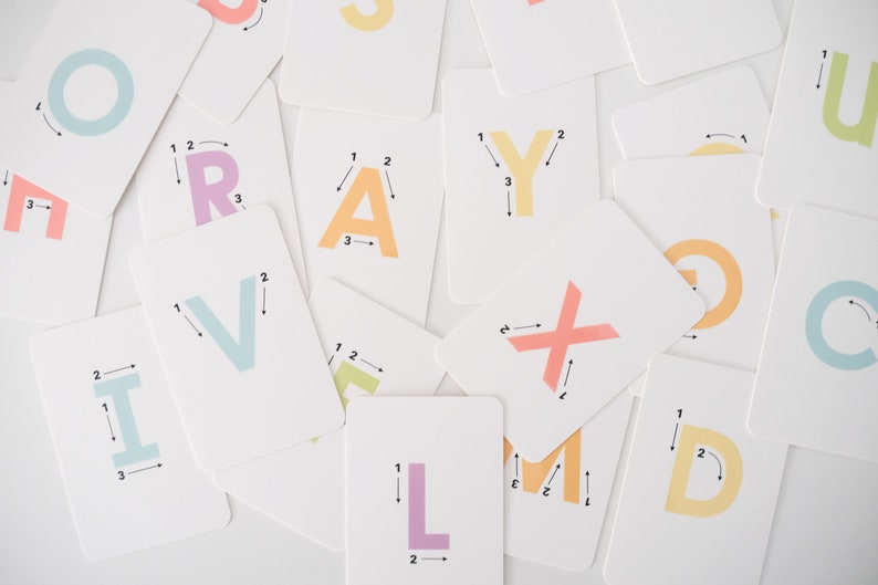 May include: A set of colorful alphabet flash cards with numbered arrows showing the stroke order for writing each letter. The cards are on a white surface.