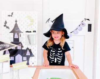 Könnte beinhalten: Ein Kind trägt einen schwarzen Hexenhut und ein schwarzes T-Shirt mit einem weißen Skelett-Druck und spielt mit einem Sinnesbecken. Das Becken ist gefüllt mit schwarzen Bohnen und grünem Gras. Das Wort "BONES" ist in weißen Buchstaben auf den schwarzen Bohnen geschrieben.