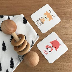 May include: A wooden baby rattle with rings and a white and black patterned bib with a black tree design. Two cards with illustrations of a deer and a fox with the words "DEER" and "FOX" printed on them.