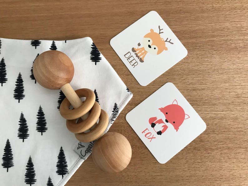 May include: A wooden baby rattle with rings and a white and black patterned bib with a black tree design. Two cards with illustrations of a deer and a fox with the words "DEER" and "FOX" printed on them.