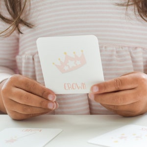 May include: A pink and gold crown card with the word "crown" printed on it. The card is being held by a person's hands. The card is part of a memory game set.