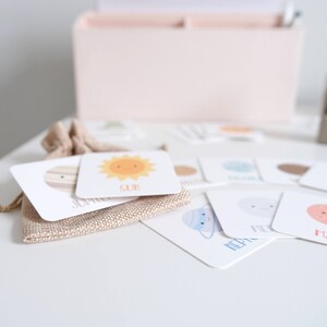 May include: A set of colorful flash cards featuring illustrations of planets and the sun. Each card has the name of the celestial body written on it. The cards are arranged on a white surface with a burlap bag in the background.