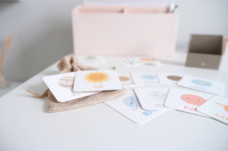 May include: A set of colorful flash cards featuring illustrations of planets and the sun. Each card has the name of the celestial body written on it. The cards are arranged on a white surface with a burlap bag in the background.