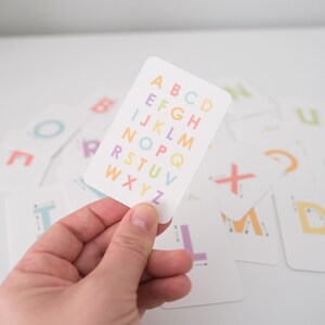 May include: A white card with the alphabet printed in rainbow colors. The letters are in a column, starting with A and ending with Z. The card is being held in a hand.