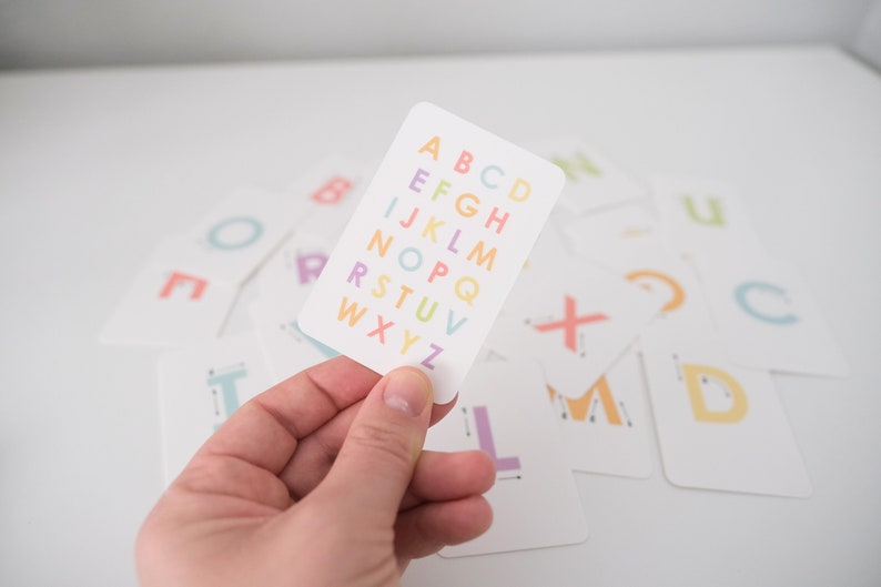 May include: A white card with the alphabet printed in rainbow colors. The letters are in a column, starting with A and ending with Z. The card is being held in a hand.