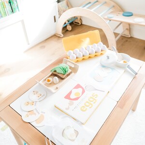 May include: A wooden table with a white surface, a yellow egg carton with white eggs, a book titled "eggs", and several small cards with images of chickens and ducks. The cards have the words "chicken", "duck", and "egg" written on them.