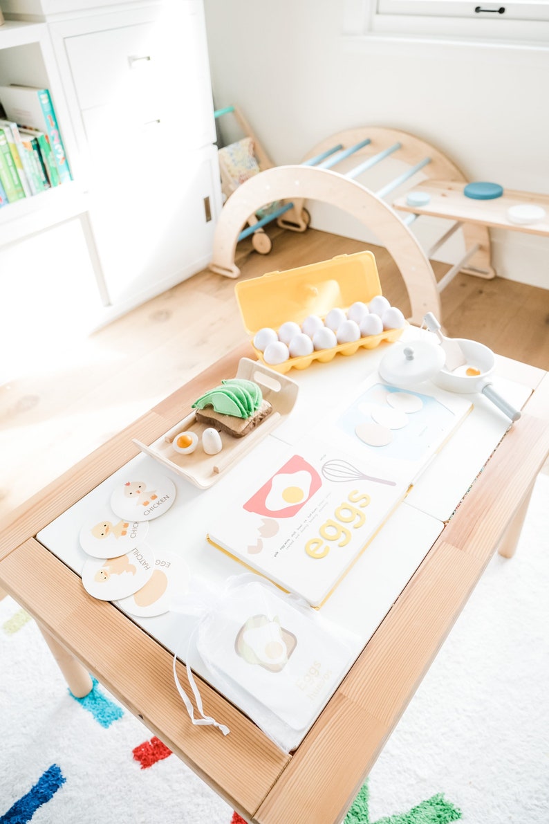 May include: A wooden table with a white surface, a yellow egg carton with white eggs, a book titled "eggs", and several small cards with images of chickens and ducks. The cards have the words "chicken", "duck", and "egg" written on them.