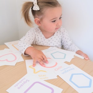 K&ouml;nnte beinhalten: Ein Kind lernt Formen mit bunten Lernkarten. Die Karten zeigen einen Stern, einen Kreis, ein Quadrat, ein Sechseck und ein Herz. Die W&ouml;rter f&uuml;r jede Form sind in Englisch und Spanisch geschrieben.