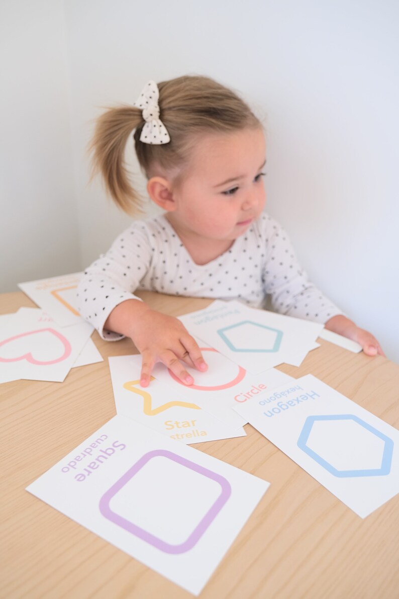 K&ouml;nnte beinhalten: Ein Kind lernt Formen mit bunten Lernkarten. Die Karten zeigen einen Stern, einen Kreis, ein Quadrat, ein Sechseck und ein Herz. Die W&ouml;rter f&uuml;r jede Form sind in Englisch und Spanisch geschrieben.