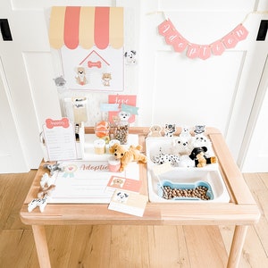 May include: A wooden play table with a white plastic insert, featuring a dog adoption theme. The table has a sign that says "Adoption Center" and a banner that says "Adoption". There are stuffed dogs, dog food, and other dog-themed accessories on the table.