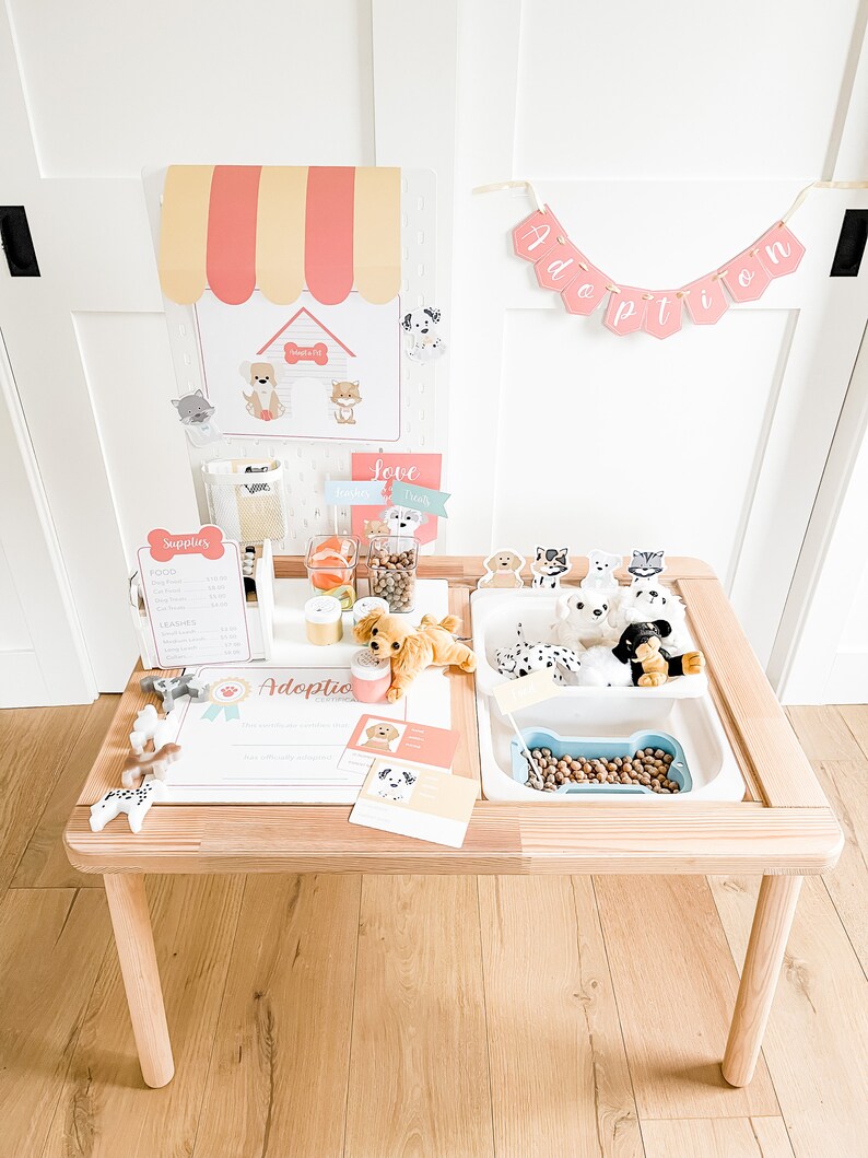 May include: A wooden play table with a white plastic insert, featuring a dog adoption theme. The table has a sign that says "Adoption Center" and a banner that says "Adoption". There are stuffed dogs, dog food, and other dog-themed accessories on the table.
