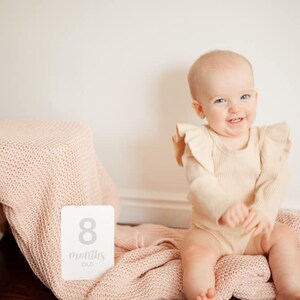 May include: A baby girl wearing a beige ruffled onesie sits on a pink knitted blanket. A white card with the text "8 months old" is in the foreground.