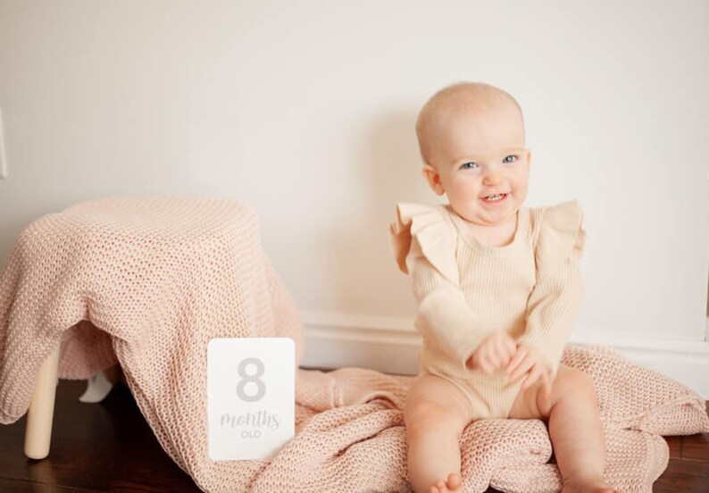 May include: A baby girl wearing a beige ruffled onesie sits on a pink knitted blanket. A white card with the text "8 months old" is in the foreground.