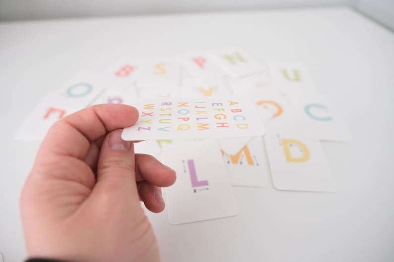 May include: A white card with the alphabet printed in rainbow colors. The letters are arranged in alphabetical order. The card is being held by a hand.