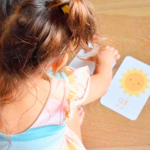 May include: A young child wearing a pink and blue striped dress with yellow hair bows plays with a flashcard featuring a yellow sun with a smiling face. The card reads "Sun is out".
