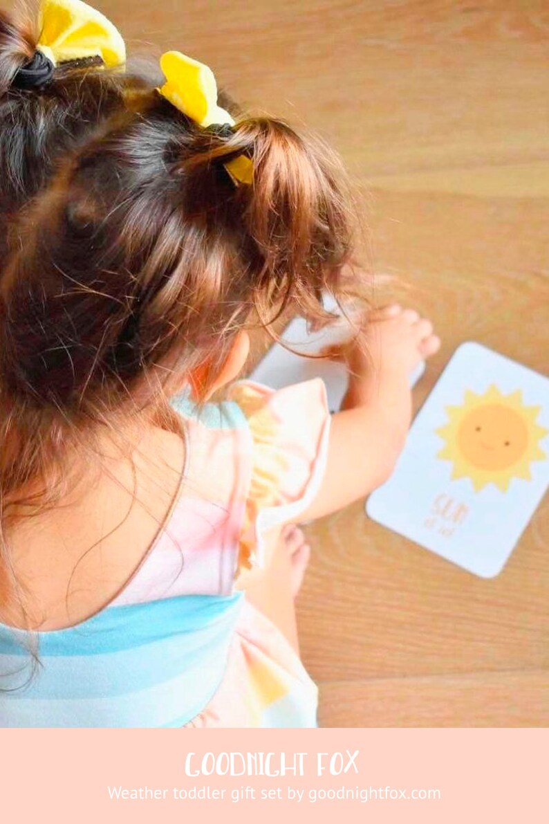 May include: A young child wearing a pink and blue striped dress with yellow hair bows plays with a flashcard featuring a yellow sun with a smiling face. The card reads "Sun is out".